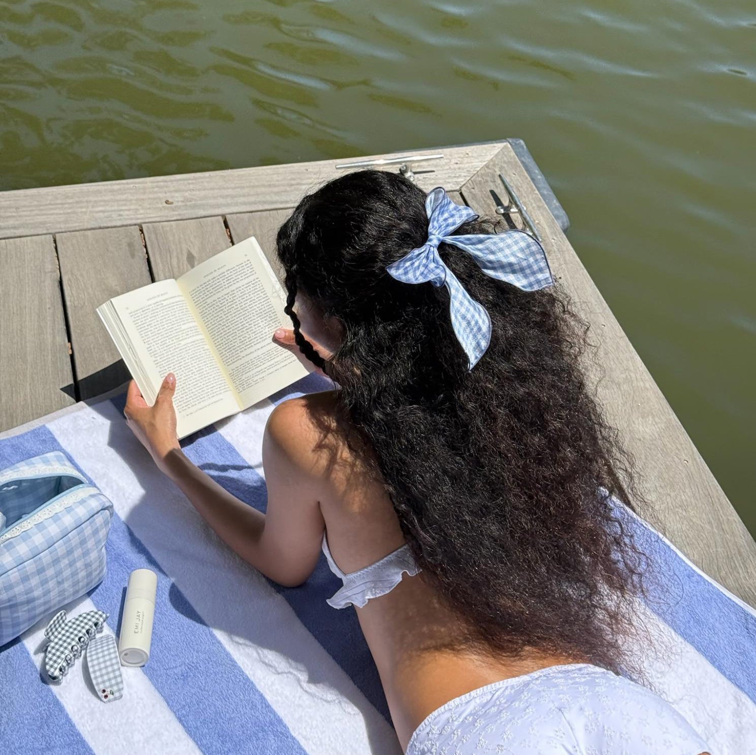 model reading a book while wearing Bow Barrette in Baby Blue Gingham in hair and laying on striped towel across wooden dock next to emi jay hair care and accessories