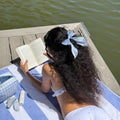 model reading a book while wearing Bow Barrette in Baby Blue Gingham in hair and laying on striped towel across wooden dock next to emi jay hair care and accessories 